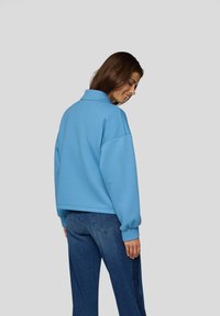 Blue textured pullover jacket with a collar and ribbed cuffs, paired with denim jeans. Back view emphasizes relaxed fit and casual design.