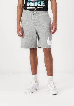 Mężczyzna ubrany w szare spodenki Nike z białym logo, białe skarpetki, białe trampki oraz czarną koszulkę Nike z grafiką, stojący na białym tle.