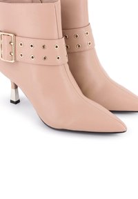 Botas de cuero nude hasta el tobillo con punta afilada, correa ancha con herrajes dorados y ojales, y acentos de tacón bloque en tono plateado.