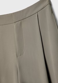 Gros plan sur un pantalon beige taille haute montrant la ceinture, un pli et une poche latérale dissimulée avec une texture de tissu lisse.