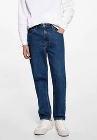 Temno modri jeans s ravnim krojem, petimi žepi in vidnimi šivi, v kombinaciji z belo kapuco in belimi supergami.
