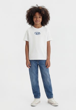 Dieťa s kučeravými vlasmi oblečené v bielom tričku s modrým logom, modrých džínsoch a bielych teniskách, stojace na jednoduchom bielom pozadí.