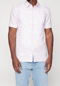 Koszula z krótkim rękawem w jasnofioletowym kolorze z kwiatowym wzorem, z kołnierzykiem zapinanym na guziki i prostym dołem, wykonana z lekkiego materiału.