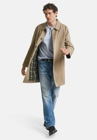 Man loopt en draagt een beige trenchcoat met een geruite voering, een lichtblauwe polo, vervaagde jeans, een zwarte riem en zwarte schoenen.