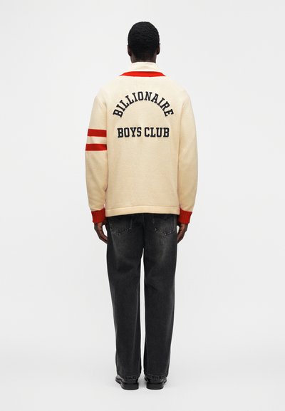Βαμβακερό μπουφάν varsity σε κρεμ απόχρωση με κόκκινες λεπτομέρειες και το κείμενο «Billionaire Boys Club» στην πλάτη. Συνδυάζεται με σκούρο τζιν και μαύρα παπούτσια.