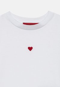 Camiseta de algodón blanca con un pequeño corazón bordado en rojo en el pecho. Cuello redondeado con una textura suave. Etiqueta de la marca visible en el interior del cuello.