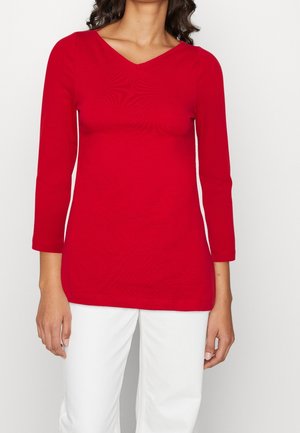 Vrouw draagt een effen rood langärmelig top met V-hals en witte broek, staand tegen een witte achtergrond.