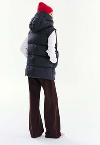 Osoba nosiaca červenú pletenú čiapku, čiernu prešívanú vestu s kapucňou, bielu blúzku s dlhým rukávom, hnedé široké nohavice a ružové topánky, stojaca bokom.