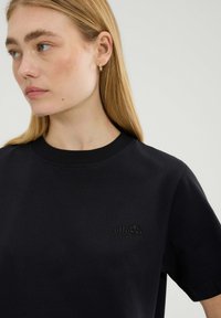 Czarny bawełniany T-shirt z okrągłym dekoltem i krótkimi rękawami; posiada wytłoczone logo na piersi oraz gładką fakturę.