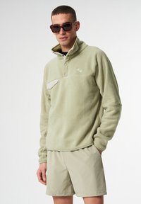 Fleece-Pullover in Hellgrün mit hohem Kragen, halber Knopfleiste und einer beigen Brusttasche. Getragen mit hellbeigen Shorts.