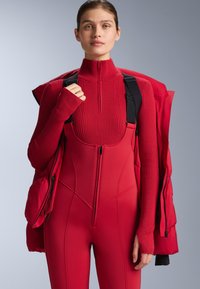 Piros termikus overál magas nyakkal, bordázott textúrával és elülső cipzárral. Piros bélelt dzsekivel párosítva, a overálon fekete pántokkal.
