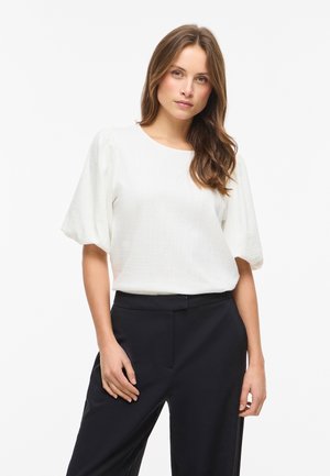 Γυναίκα με μακριά καστανά μαλλιά που φοράει μια λευκή υφασμάτινη μπλούζα με κοντά μανίκια και μαύρο παντελόνι, στέκεται μπροστά σε λευκό φόντο.