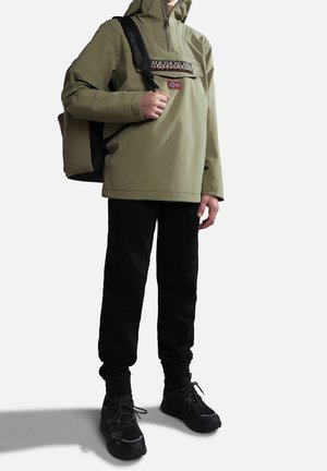 Veste imperméable verte avec un col zippé, patch logo, pantalons noirs et baskets noires à lacets. Sac à dos avec une bandoulière.