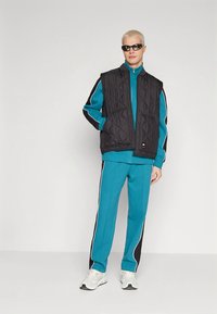 Juodas siuvinėtas liemenė ant mėlynos sportinės aprangos su juodais ir baltais akcentais. Turi aukštą apykaklę, užtraukiamą priekinę dalį ir sportinius sportbačius.