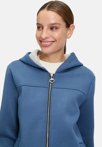 Niebieska bluza z zamkiem błyskawicznym, wykonana z miękkiego materiału, z szarym wnętrzem, z kapturem, kieszeniami z przodu oraz srebrnym detalem zamka.