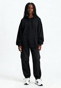 Čierna oversized mikina s prednou vreckom, šnúrkami a rebrovanými manžetami, skombinovaná s čiernymi cargo nohavicami a bielymi teniskami.