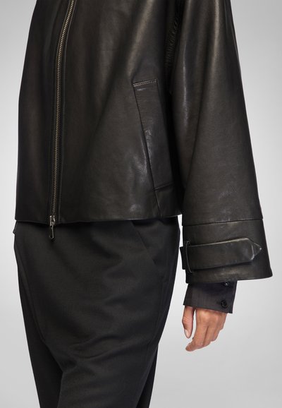 Black leather jacket with a slight sheen, featuring a stand-up collar, zip closure, and a fitted design. The sleeve has a strap detail.