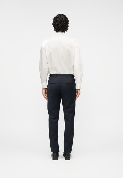 Άσπρο πουκάμισο με μακριά μανίκια συνδυασμένο με μπλε σκούρο παντελόνι. Το παντελόνι διαθέτει δύο πίσω τσέπες και στενή γραμμή. Μαύρα παπούτσια ολοκληρώνουν την εμφάνιση.