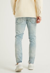 Persona con jeans azul claro, zapatillas blancas y chaqueta de forro polar beige, de pie contra un fondo blanco liso, de espaldas.