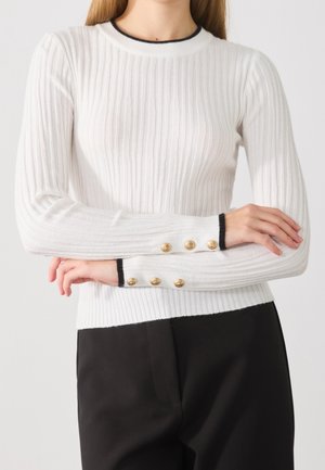 Personne portant un pull blanc à manches longues côtelé avec des bordures noires et des boutons dorés aux poignets, associé à un pantalon noir.