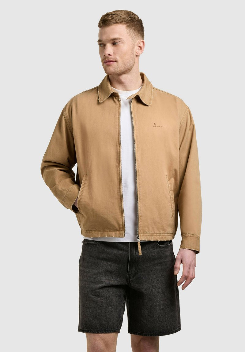 Homme portant une veste beige à fermeture éclair sur une chemise blanche et un short en jean noir, debout avec une main dans la poche, regardant sur le côté.