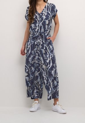 Kvinde iført en marineblå og hvid trykt jumpsuit med korte ærmer og hvide sneakers, poserende med den ene hånd i lommen mod en ensfarvet baggrund.