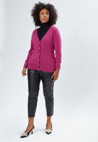 Cardigan fuchsia avec un col en V, des manches longues et des détails en bouton sur un collant noir, associé à un pantalon en simili cuir noir et des talons décorés.