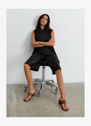 Hosszú sötét hajú nő egy fekete irodai székben ül, fekete ujjatlan felsőt, térdig érő fekete rövidnadrágot és barna magas sarkú szandált visel.