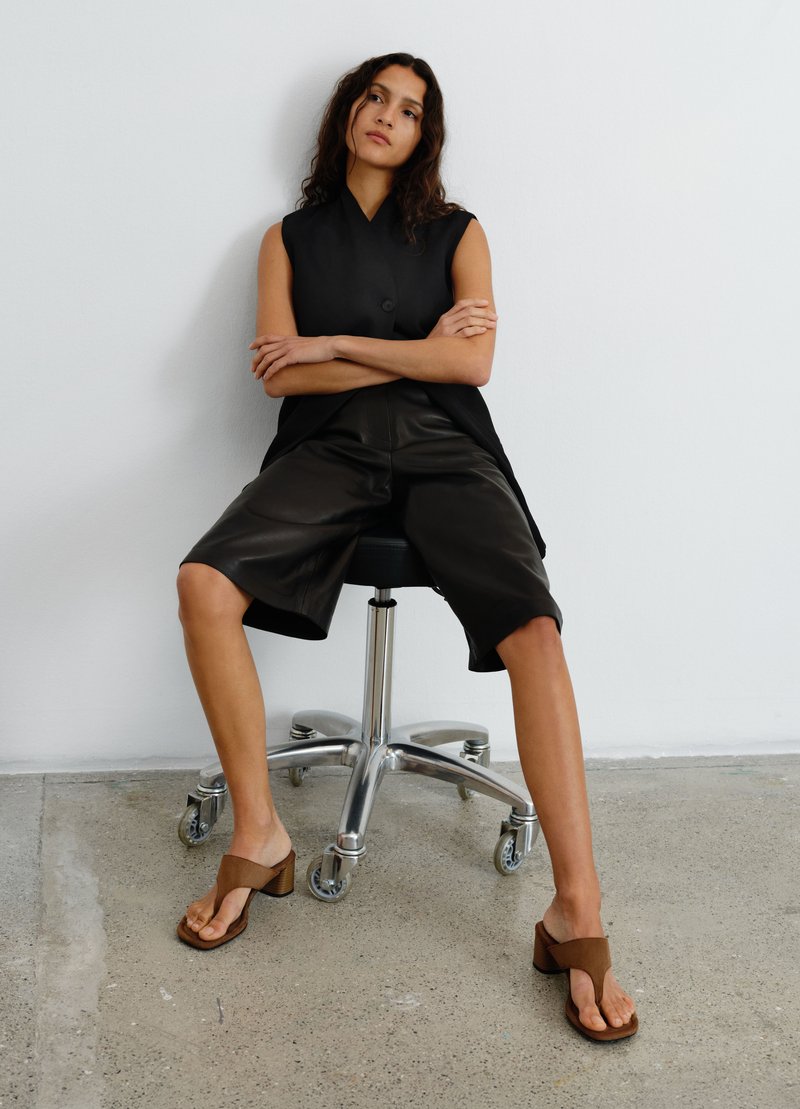 Vrouw met lang donker haar zit op een zwarte bureaustoel, draagt een zwart mouwloos topje, zwarte shorts tot op de knie en bruine sandalen met hak.