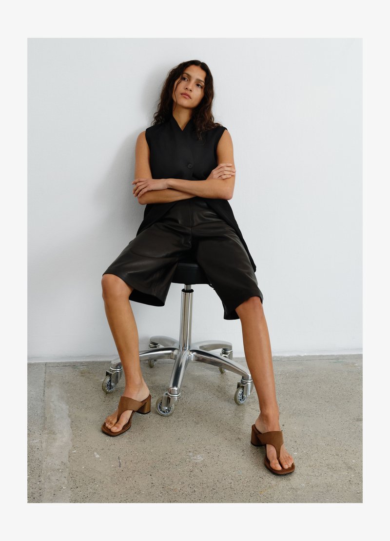 Vrouw met lang donker haar zit op een zwarte bureaustoel, draagt een zwart mouwloos topje, zwarte shorts tot op de knie en bruine sandalen met hak.