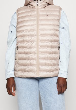 Beige gesteppte ärmellose Daunenweste mit frontalem Reißverschluss, getragen über einer hellblauen Jeansjacke und Jeans.