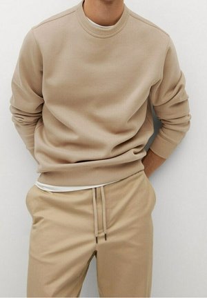 Mann trägt beigen Pullover und passende beige Jogginghose, Hände in den Taschen, steht vor einem schlichten, hellen Hintergrund.