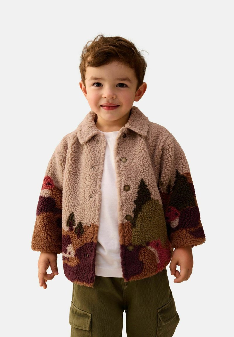 Plush, beige shearling jacket with colorful mountain patterns, button front, and notched collar. Paired with a white shirt and olive cargo pants.