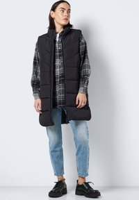 Svart ermeløs puffervest med høy krage, quiltet mønster, kombinert med rutete skjorte og lyseblå jeans, brukt med svarte sko.