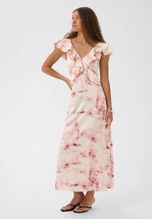 Femme aux cheveux bouclés portant une robe maxi à volants rose et crème et des sandales noires, debout avec une main sur la hanche, regardant vers la gauche.