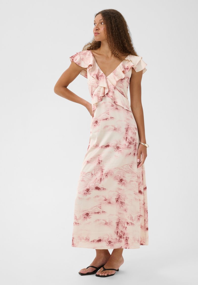 Femme aux cheveux bouclés portant une robe maxi à volants rose et crème et des sandales noires, debout avec une main sur la hanche, regardant vers la gauche.