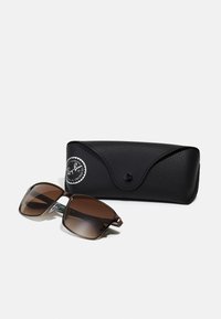 Ochelari de soare căprui cu lentile pătrate, ramă din metal și accente din plastic, însoțiți de o husă texturată neagră cu un logo.