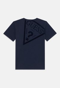 T-shirt bleu marine avec un grand imprimé foncé du nom de la marque "GUESS" en forme triangulaire et un texte supplémentaire au dos.