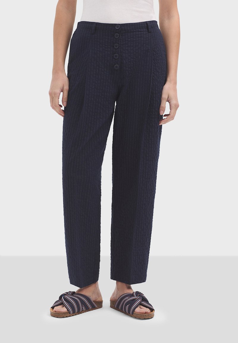 Tamno plave hlače s teksturom i vertikalnim prugama, visoki struk i zatvaranje na pet dugmadi, u kombinaciji s natikačama na prugice.