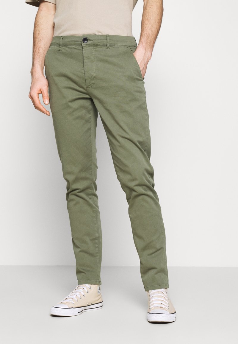 Homme portant un pantalon slim vert olive avec les mains dans les poches et des baskets montantes beiges, debout devant un fond uni.