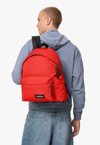 Mochila Eastpak roja con un diseño redondeado, cremalleras negras, bolsillo frontal y correas ajustables, llevada por una persona con una sudadera gris.