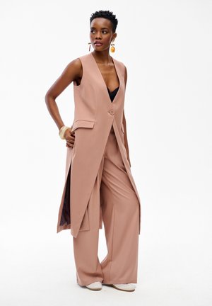 Kobieta o krótkich włosach, ubrana w beżową, bezrękawnikową, długą kamizelkę, dopasowane szerokie spodnie, czarną bluzkę, białe buty oraz duże kolczyki, pozująca na stojąco.