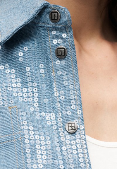Ανοιχτό μπλε τζιν μπουφάν με λευκές κυκλικές παγιέτες και μεταλλικά κουμπιά με σχέδιο σταυρού, φορεμένο πάνω από λευκό τοπ.