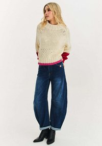 Bílý pletený svetr s volným střihem a růžovo-červenými strukturovanými manžetami, kombinovaný s tmavě modrými širokými džíny a černými špičatými kotníkovými botami.