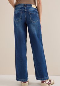 Jean en denim bleu à jambes larges avec une texture délavée, deux poches arrière et des coutures contrastantes. Détail minimaliste de la ceinture visible.