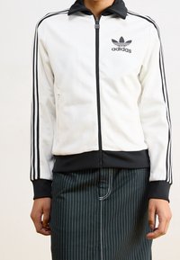 Giacca sportiva bianca con dettagli neri, chiusura con zip e tre strisce nere sulle maniche. Logo Adidas sul petto sinistro. Tessuto liscio.