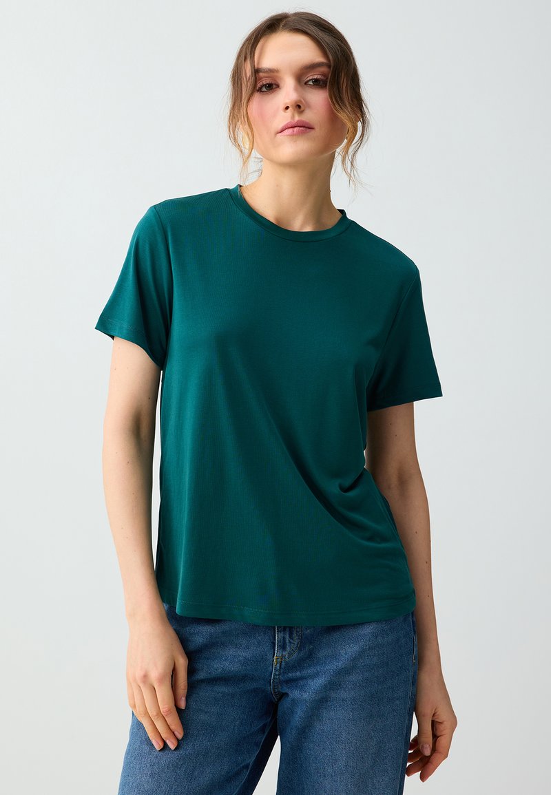 Jeune femme aux cheveux châtain clair attachés porte un t-shirt vert foncé à manches courtes et un jean bleu, debout devant un fond uni.