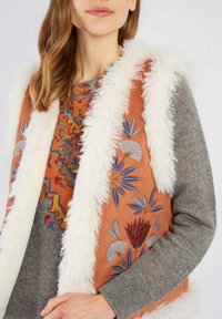 Brązowa kamizelka z sztucznego futra z białym wykończeniem i kolorowym haftem w kwiaty. Pod nią znajduje się wzorzysty sweter w odcieniach szarości.