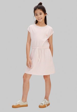 Ragazza giovane con capelli scuri raccolti in uno chignon, che indossa un vestito a righe rosa e bianche e scarpe da ginnastica beige, in piedi con una mano in tasca su sfondo grigio.