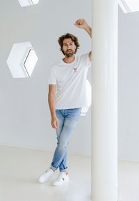 T-shirt bianca con una piccola grafica rossa e blu, jeans azzurri chiari e sneakers bianche con dettagli verdi, in piedi contro una colonna bianca.